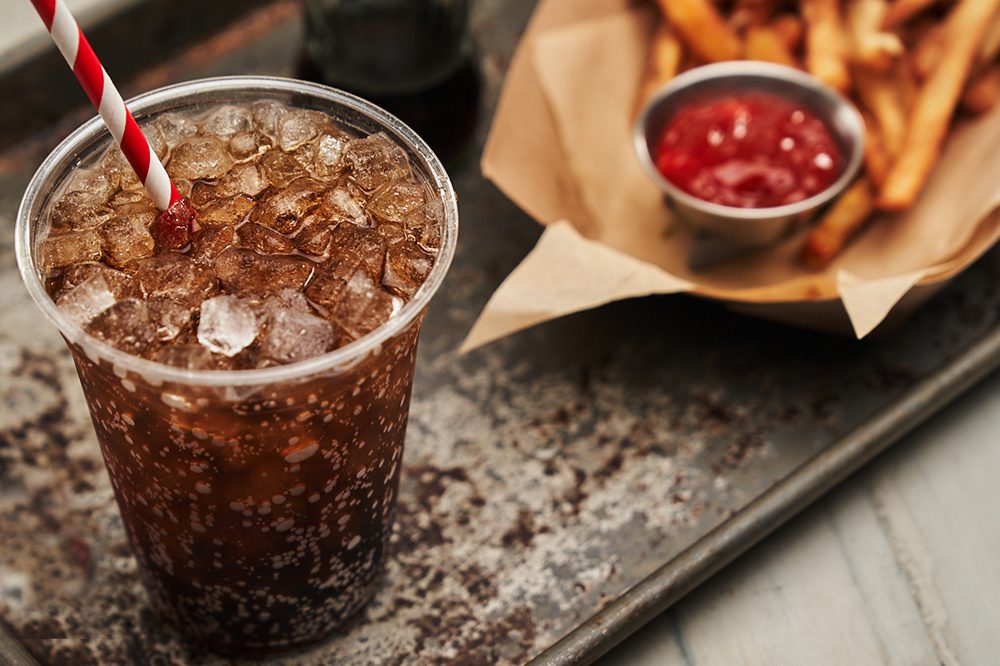 A cola drink with cubelet ice sits on a counter next to some fried and ketchup.