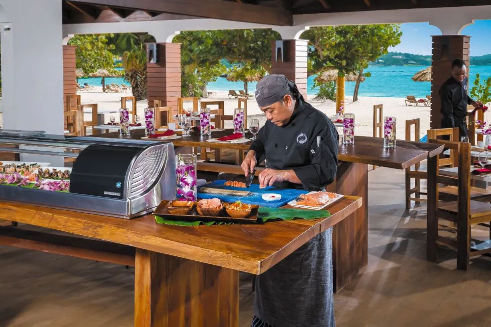 A sushi chef in a luxury outdoor kitchen at a resort is preparing food.