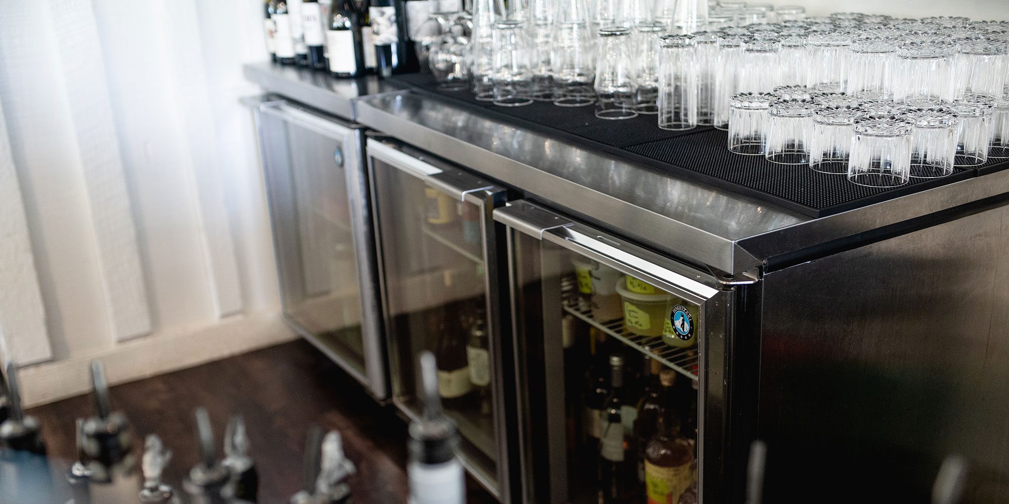 A bar cart with glasses on top and numerous bottles inside a chilled cabinet.