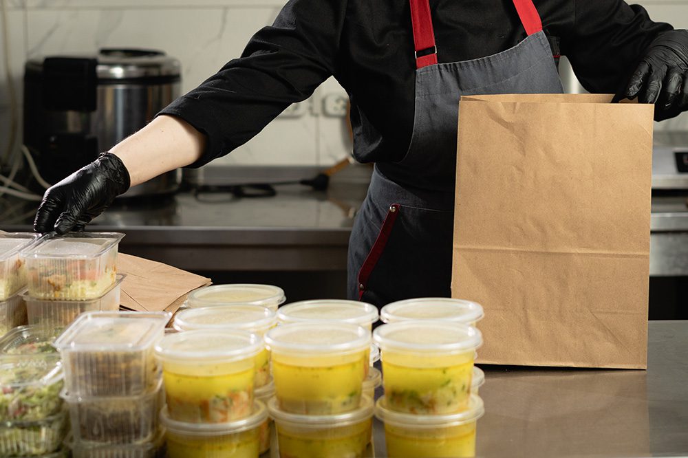 Worker packing hot food containers into a paper bag in a commercial kitchen, illustrating streamlined order staging with Hoshizaki pass-thru cabinets.
