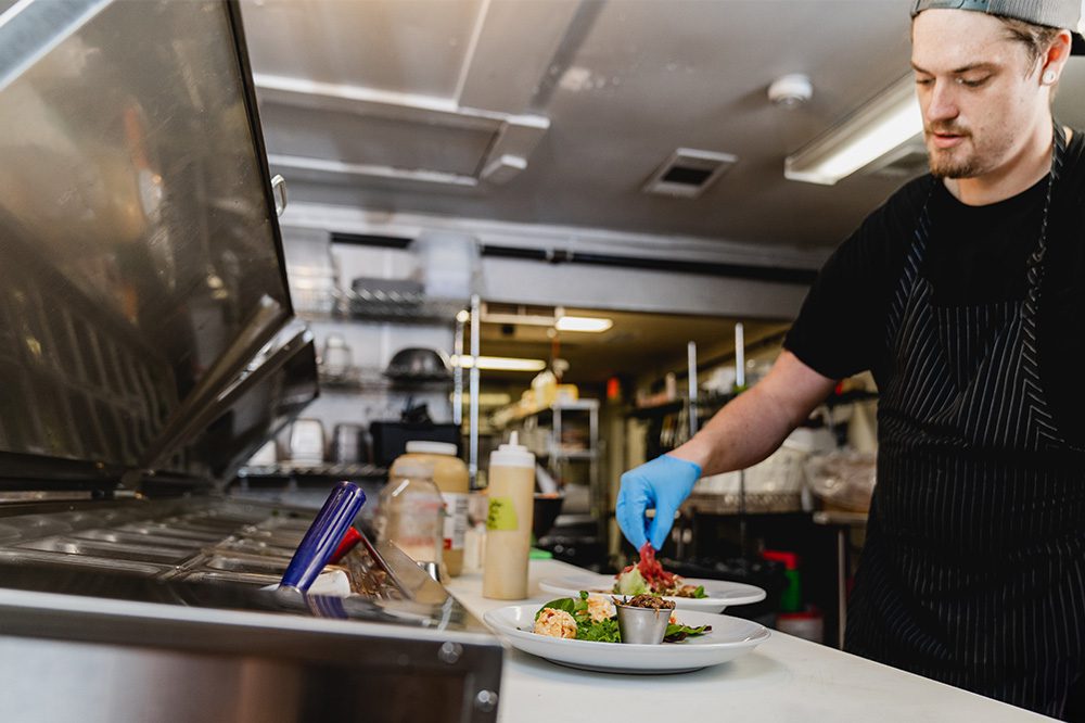 Chef preparing plates in a busy kitchen using a Hoshizaki sandwich prep table with stainless steel food storage and prep surface.