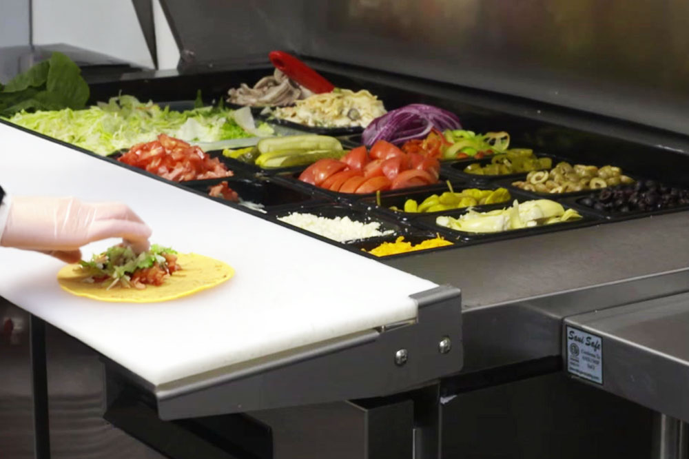 A person prepares a taco on a counter next to a salad bar with various fresh vegetables and toppings in trays.