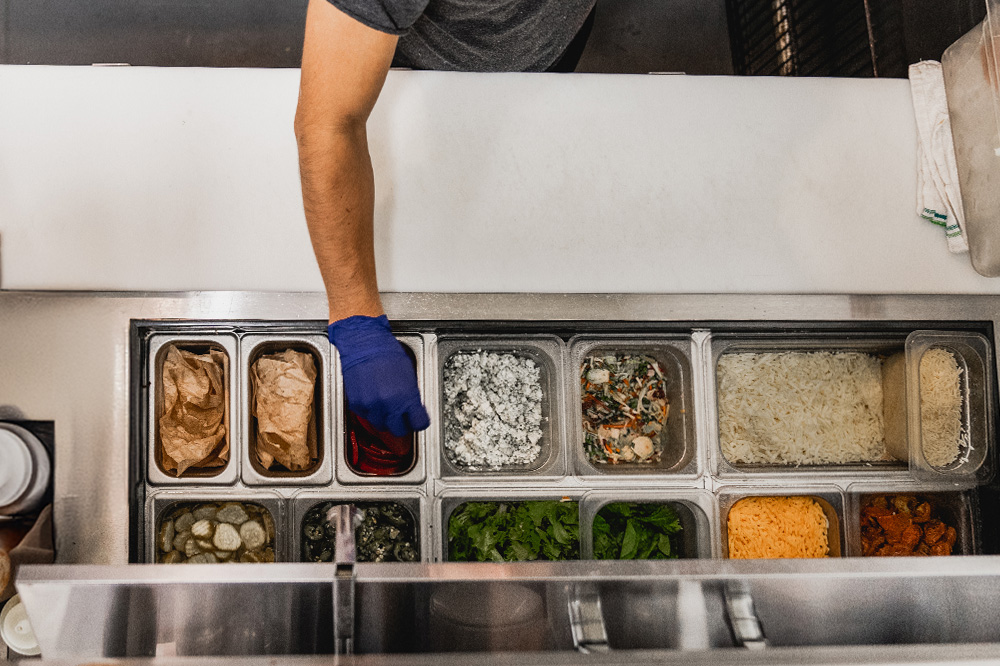 Person with a blue glove reaching into a container in a food prep station with various ingredients, viewed from above.