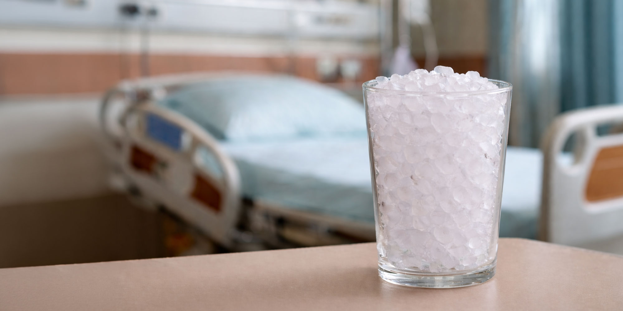 A glass filled with ice chips sits on a table in front of an out-of-focus hospital bed.