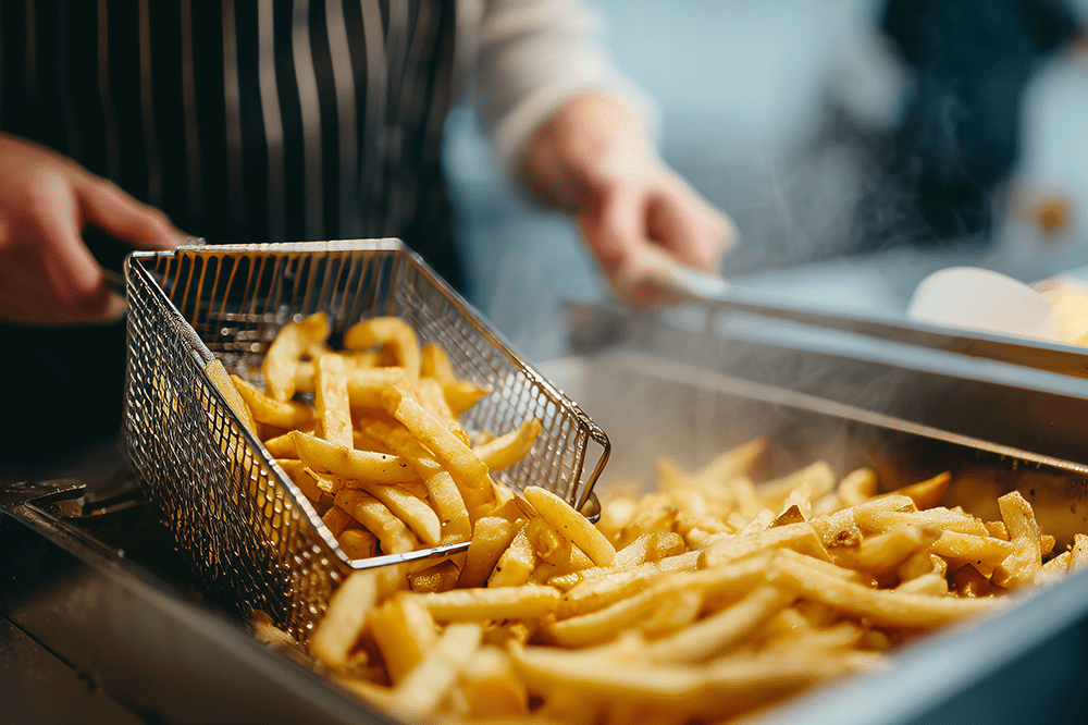 A person in a striped apron lifts a basket of freshly cooked French fries from a deep fryer, with more fries visible below.