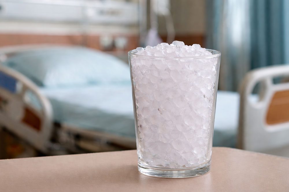 A clear glass filled with ice chips sits on a table in a hospital room with a hospital bed in the background.