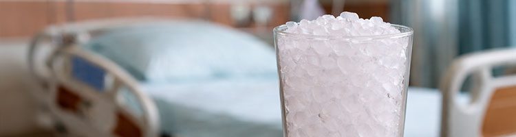 A clear glass filled with ice chips sits on a table in front of a hospital bed in a patient room.