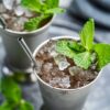 Two silver cups filled with crushed ice and garnished with fresh mint sprigs, each with a metal straw, sit on a gray surface.