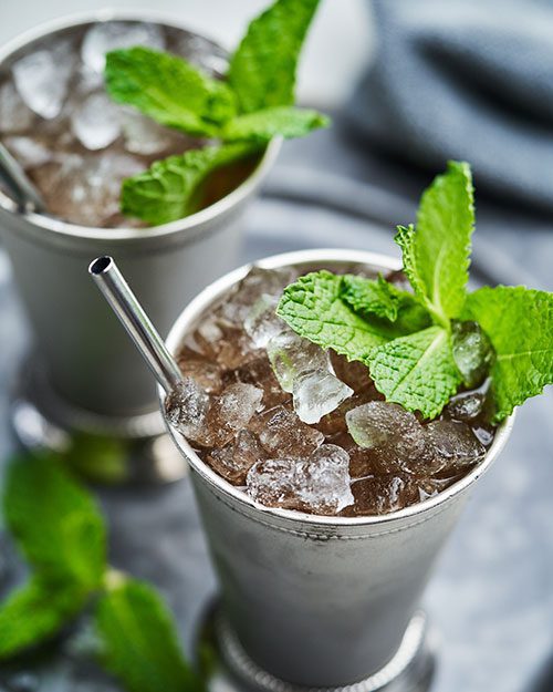 Two silver cups filled with crushed ice and garnished with fresh mint sprigs, each with a metal straw, sit on a gray surface.