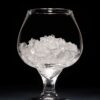 A clear glass snifter filled with crushed ice sits on a dark surface against a black background.