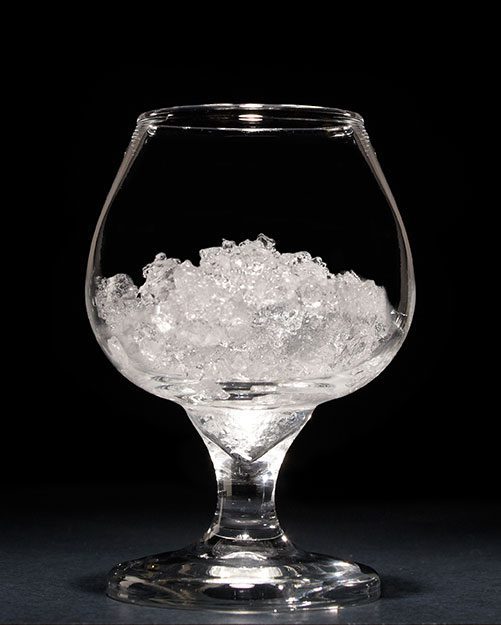 A clear glass snifter filled with crushed ice sits on a dark surface against a black background.