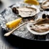 Three opened oysters on a bed of ice with lemon wedges and a small fork on a dark plate.