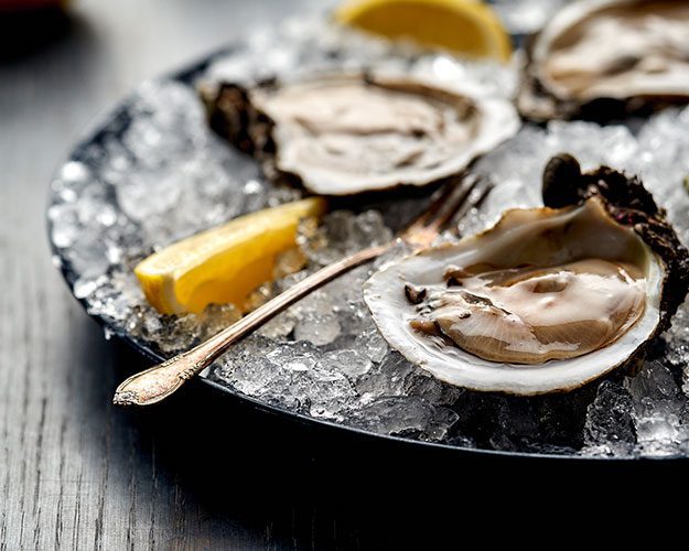 Three opened oysters on a bed of ice with lemon wedges and a small fork on a dark plate.