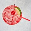 A red cocktail with crushed ice, a red straw, garnished with a cherry and a lime wedge, seen from above on a bed of ice.
