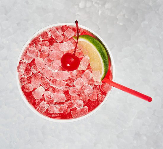 A red cocktail with crushed ice, a red straw, garnished with a cherry and a lime wedge, seen from above on a bed of ice.