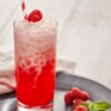 A tall glass of red and white layered iced drink with a striped straw, topped with a raspberry, sits on a plate with raspberries and mint beside it.