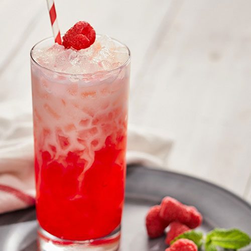 A tall glass of red and white layered iced drink with a striped straw, topped with a raspberry, sits on a plate with raspberries and mint beside it.