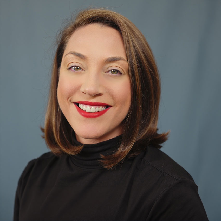 Woman with shoulder-length brown hair, wearing a black turtleneck, smiles while facing the camera against a plain blue background.