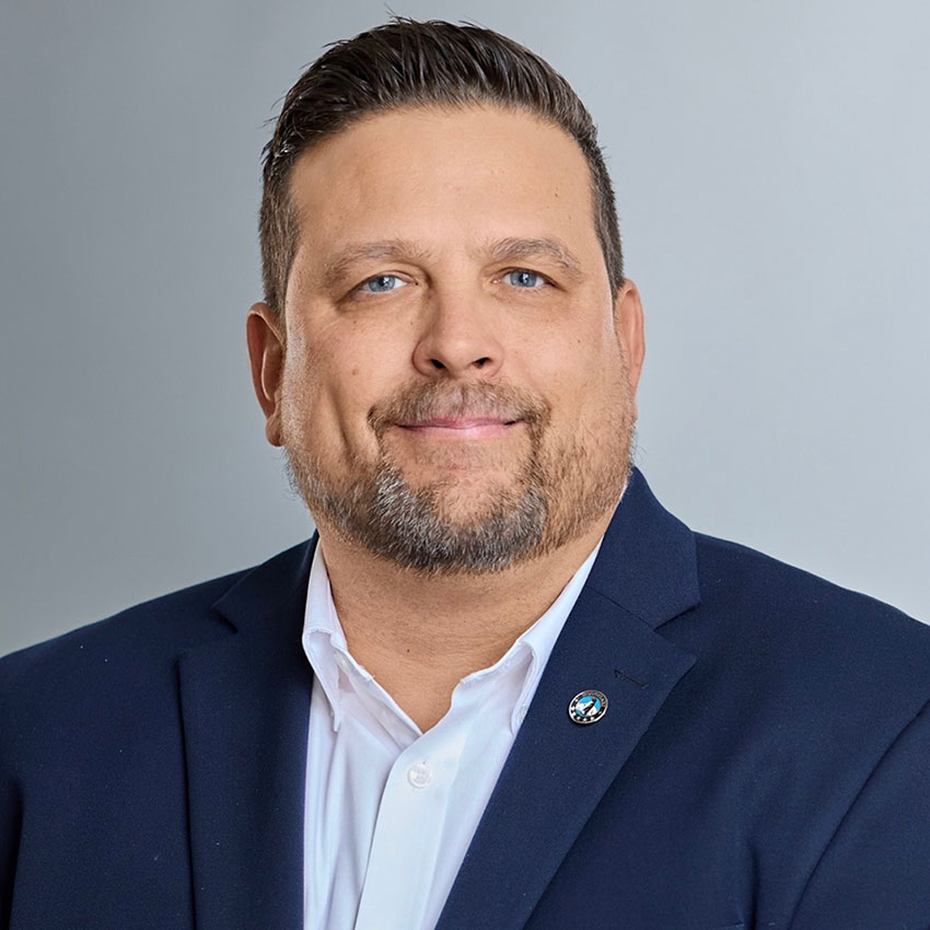 Man with short brown hair and beard wearing a dark suit jacket, white shirt, and a pin, posing in front of a plain light gray background.