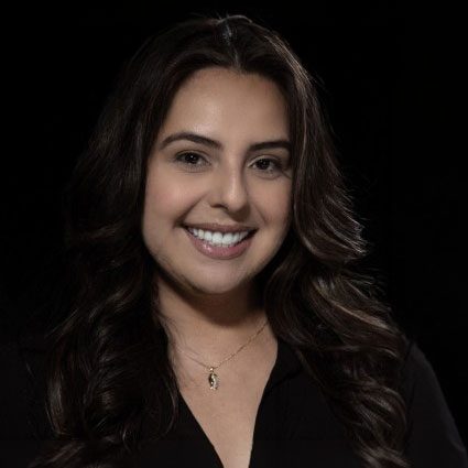 Woman with long dark hair, wearing a black top and a necklace, smiling at the camera against a dark background.