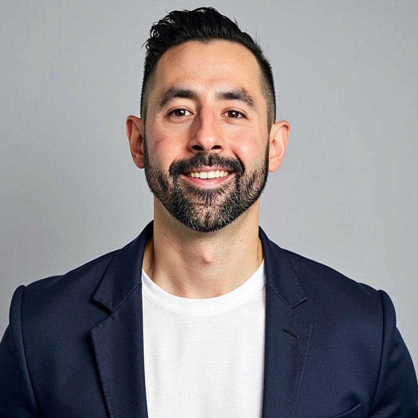 Man with short dark hair and beard wearing a navy blazer over a white t-shirt, smiling in front of a plain gray background.