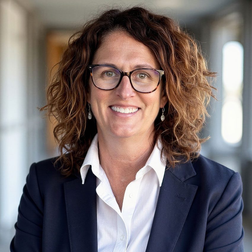 Woman with curly brown hair and glasses, wearing a navy blazer and white shirt, smiling, standing in a hallway with blurred background.